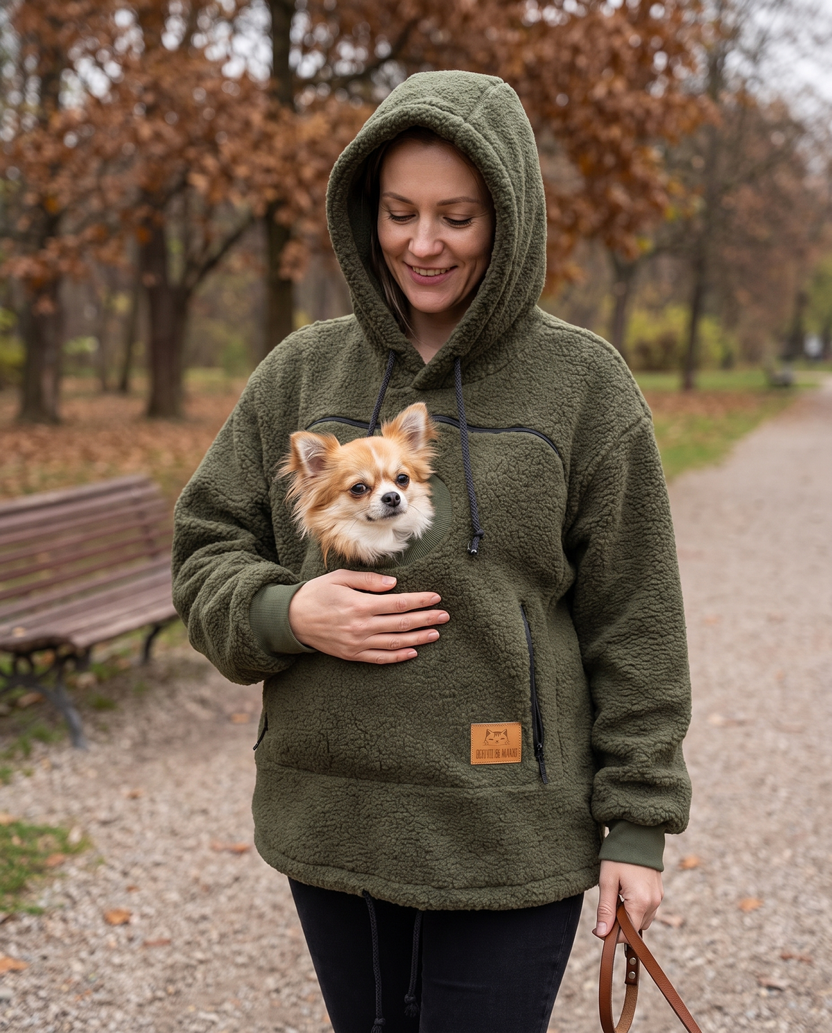 Woman in an army green carrier hoodie walking in an autumn park with a small dog resting in the front pouch