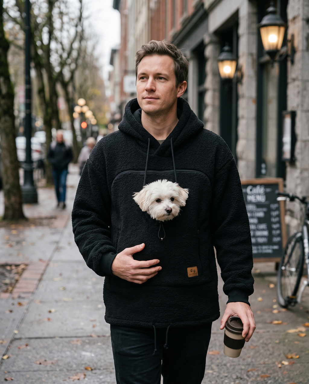 Man walking in a black carrier hoodie with a small white dog looking out of the front opening